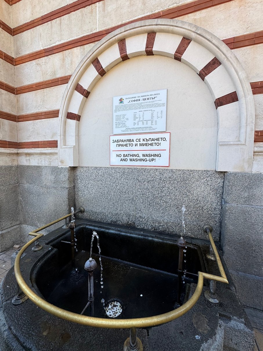 Warm mineral water flows from a public tap in a striped stone niche in central Sofia.