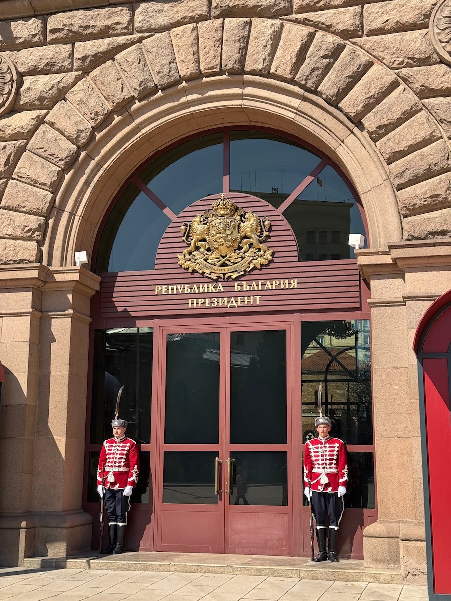 Two guards in red uniforms stand at the arched entrance of the Presidency building in Sofia.