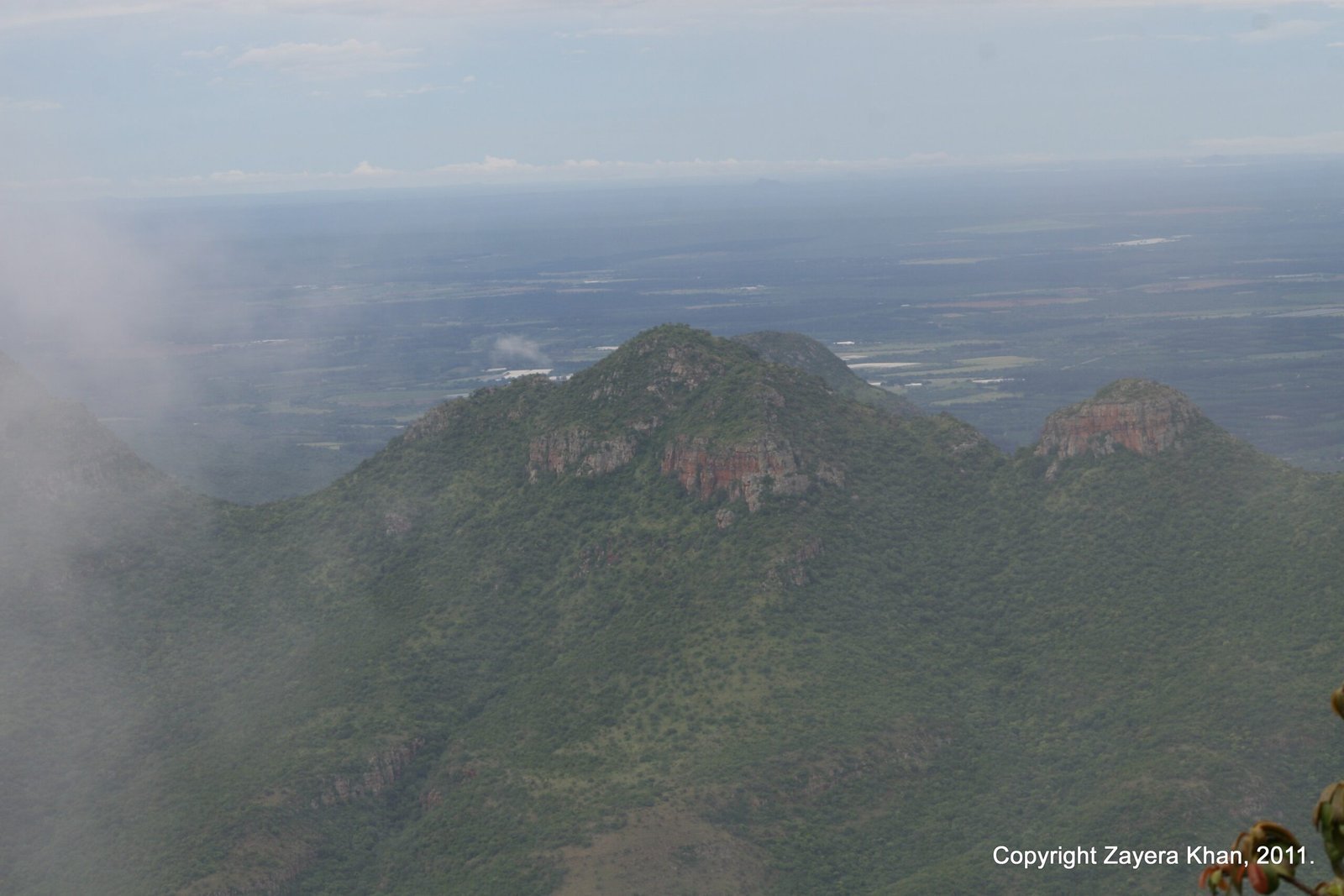 Green mountains and a wide plain show below clouds from the air over South Africa.