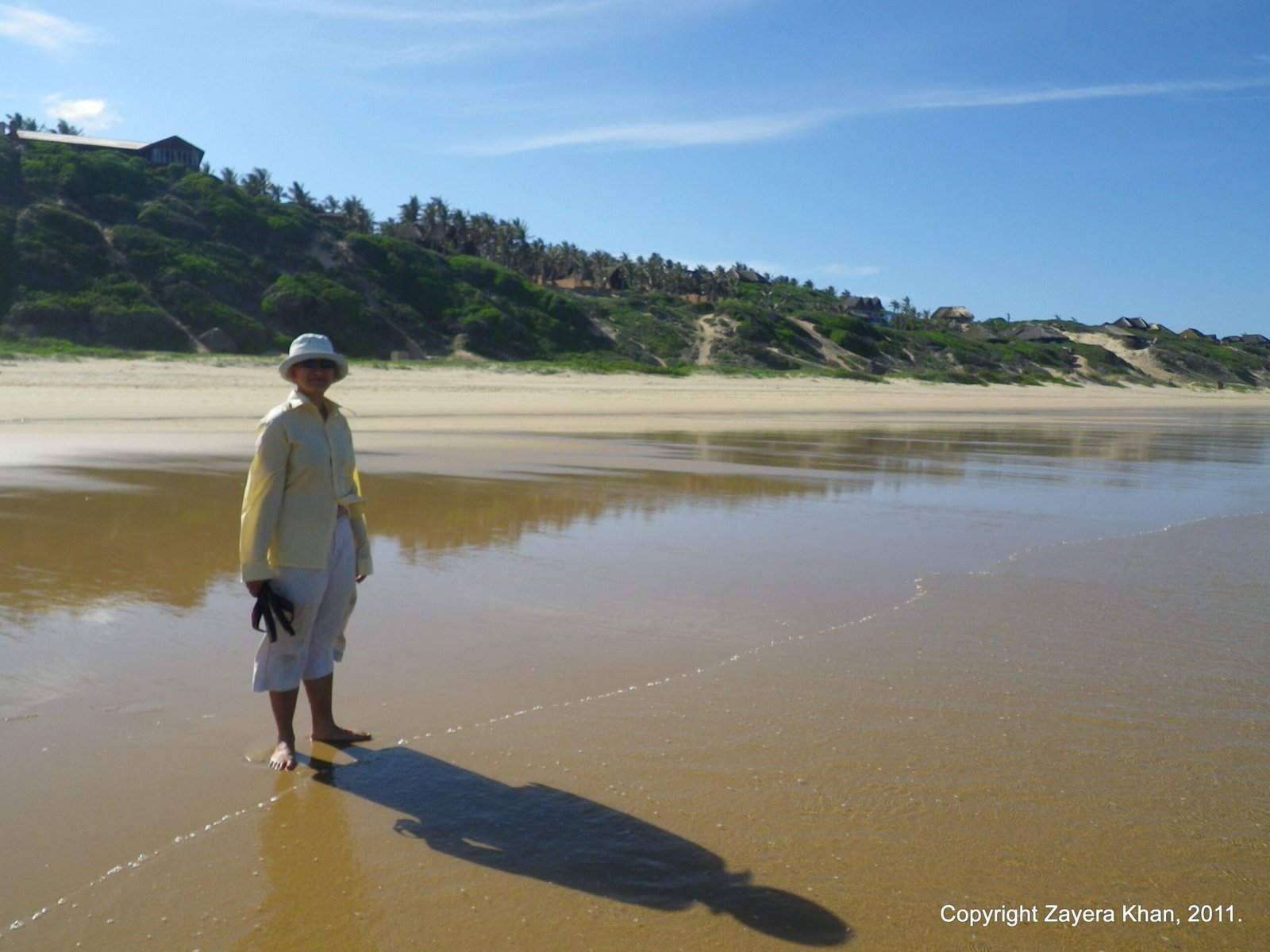 Zayera Khan stands barefoot on a wide wet beach below green dunes in southern Africa.