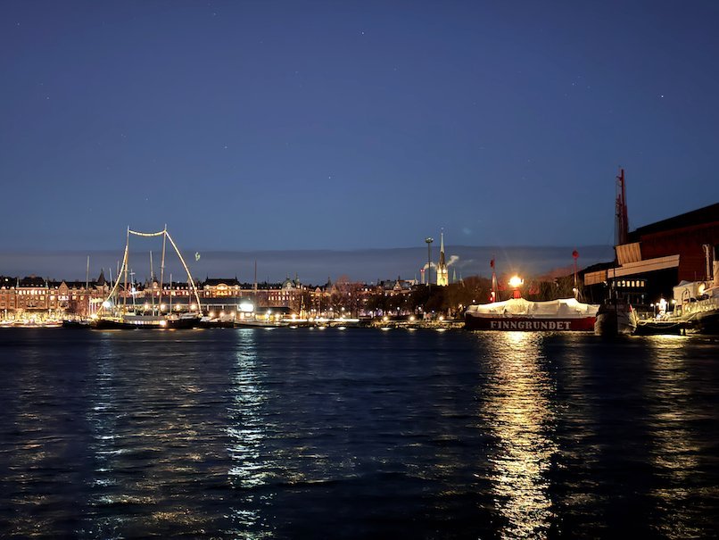 Stockholm waterfront at dusk, with tall ship masts and city lights reflected in the water
