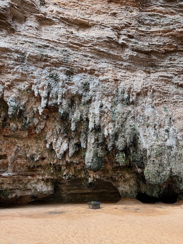 White mineral formations hang from a rock overhang above sand.