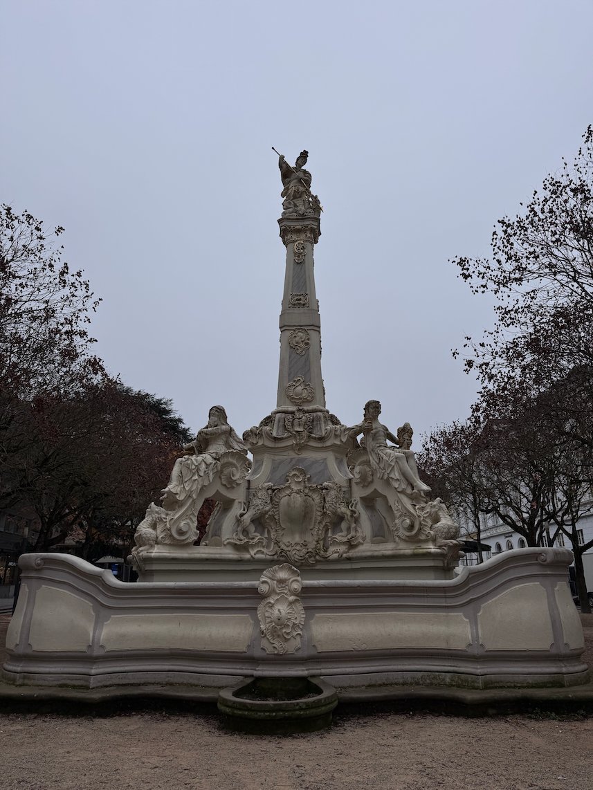 A baroque fountain with a tall column and statues stands on a square in Trier.