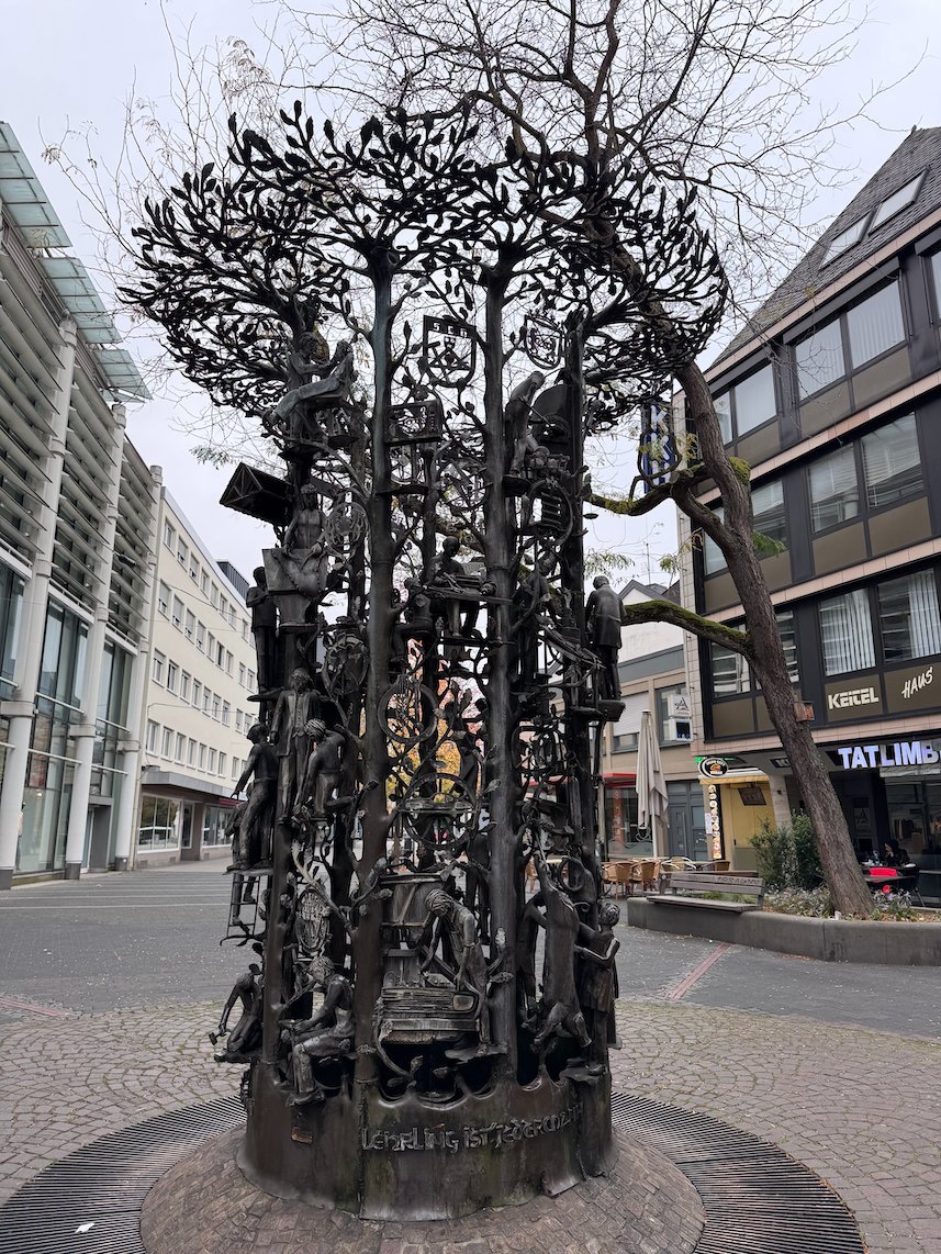 A bronze sculpture with trees and figures stands next to a bare tree in Trier.