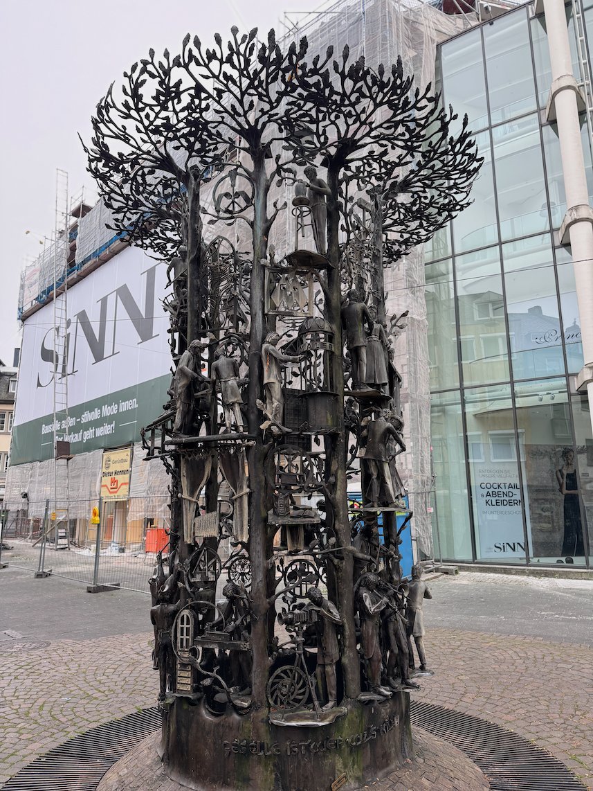 A bronze sculpture with many small figures stands in a shopping street in Trier.