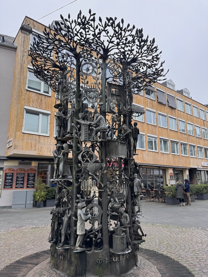 A tall bronze sculpture shaped like a tree with figures stands on a square in Trier.