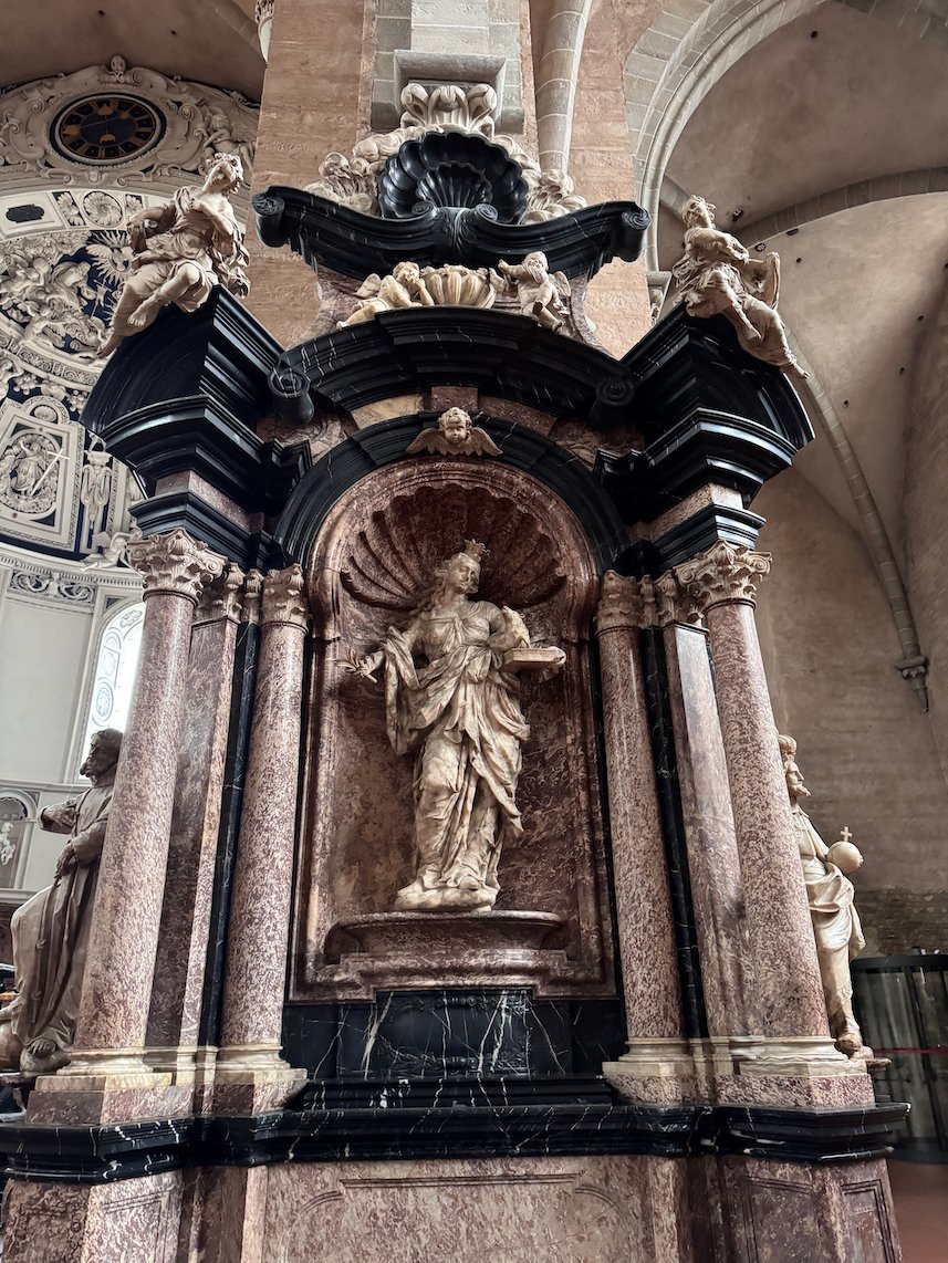 A white statue of a saint stands in a shell niche of a baroque marble altar in Trier Cathedral.