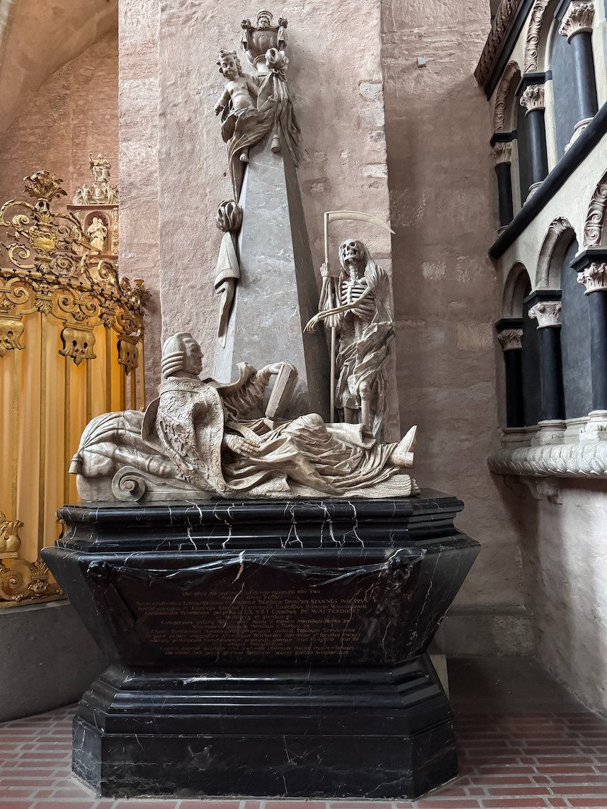 A marble tomb with carved figures stands next to a pillar in Trier Cathedral.