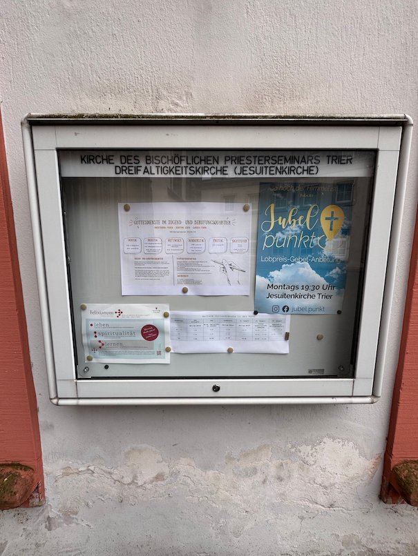 A glass notice board with church information hangs on a wall in Trier.