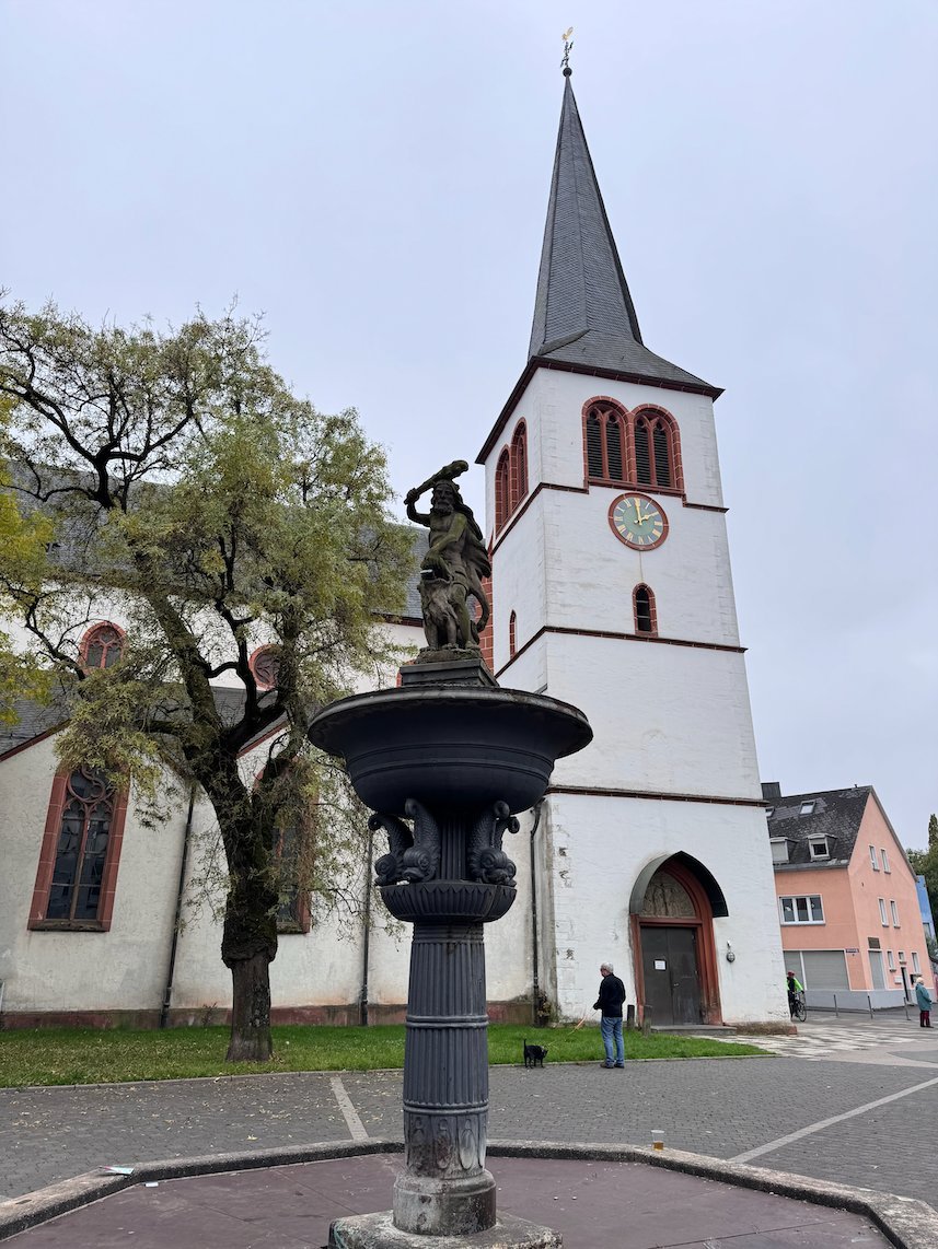 A church with a white tower stands behind a small fountain in Trier.