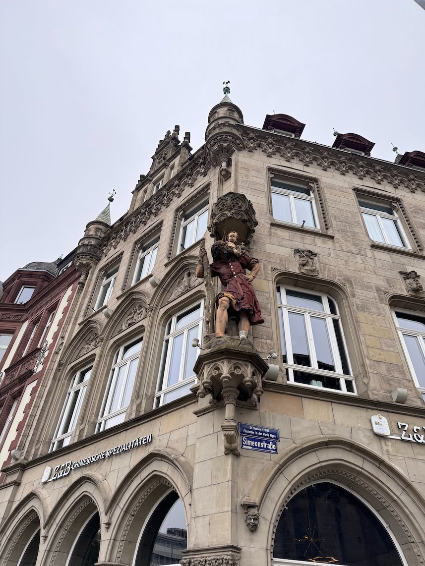 A painted statue stands on the corner of an ornate sandstone building in Trier.