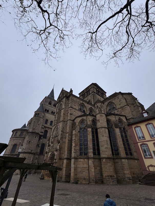 A Gothic church stands under bare tree branches on a square in Trier.