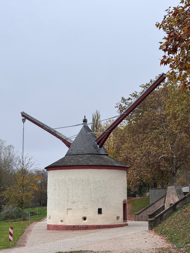 A round white crane house with a wooden arm stands next to the Moselle in Trier.