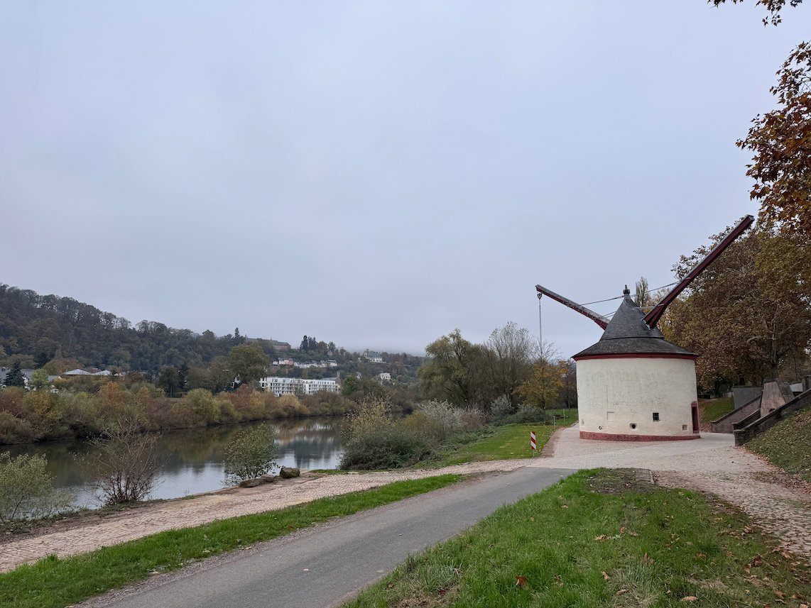 A path runs along the Moselle riverbank toward an old stone crane in Trier.