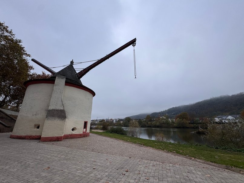 A historic stone crane with a wooden arm stands on the bank of the Moselle in Trier.