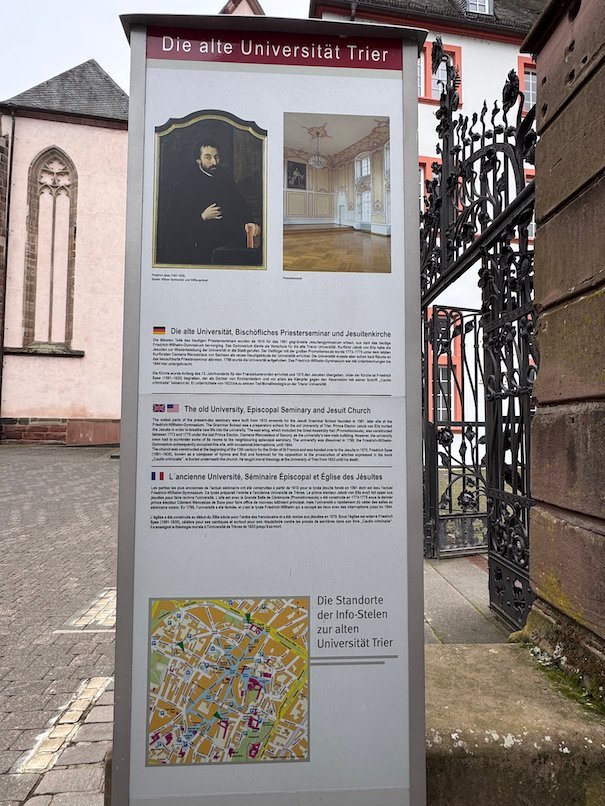 An information sign about the old University of Trier stands by an iron gate.