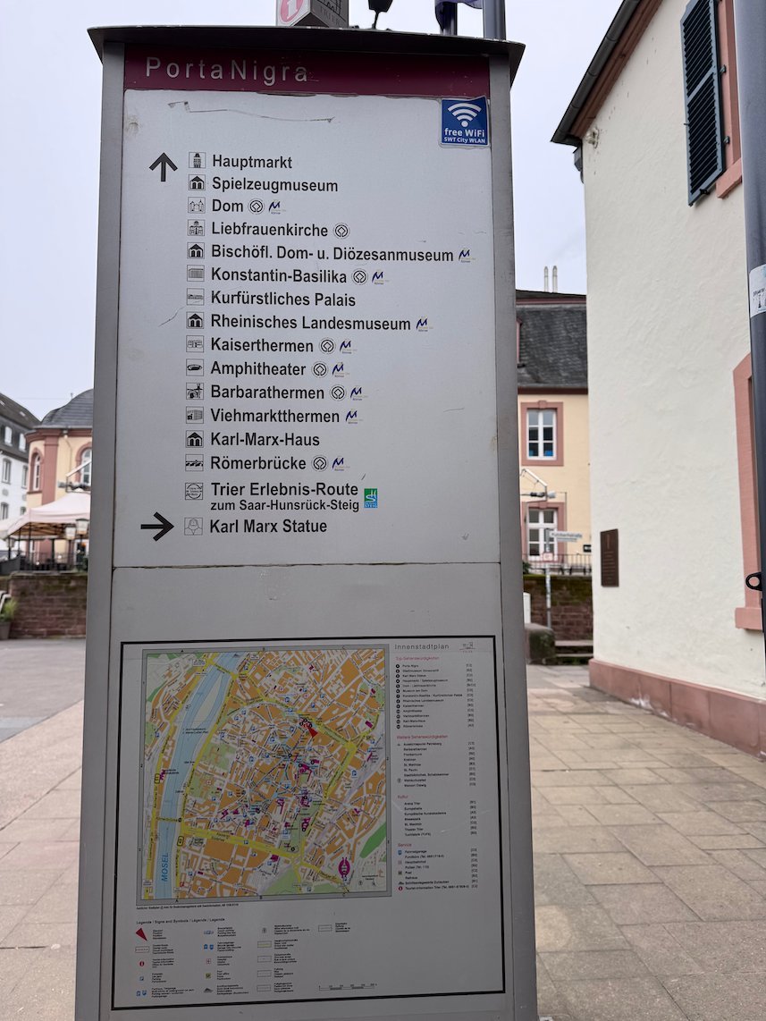 A signpost with a city map and directions stands near the Porta Nigra in Trier.