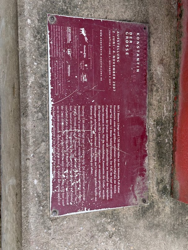 A red plaque with text hangs on a stone wall in Trier.