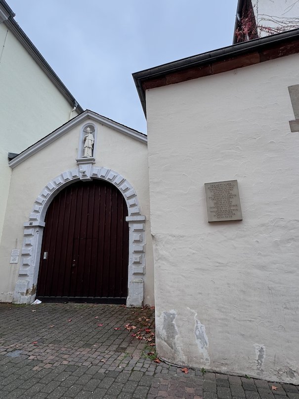 A white chapel with a red arched door stands on a cobbled street in Trier.