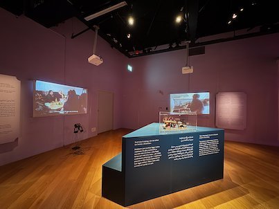 Dark exhibition room with screens on the walls and a lit glass showcase
