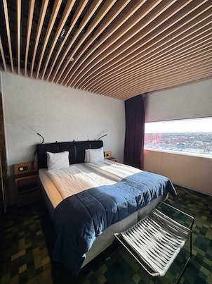 Hotel bed with a blue cover, a slatted wooden ceiling and a wide window view over Umeå