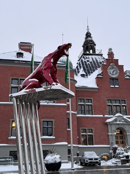Red animal sculpture on top of steel poles in front of a historic red brick building in Umeå