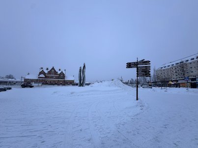 Snowy square in Umeå with a signpost and the red brick railway station in the distance, on a grey winter day