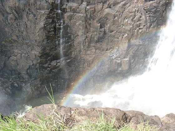 A rainbow shines in front of a dark rock wall and falling water at Victoria Falls.