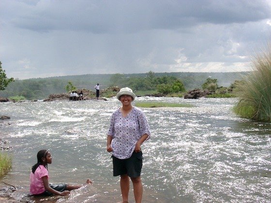 A traveler in a hat stands in the shallow Zambezi River above Victoria Falls.