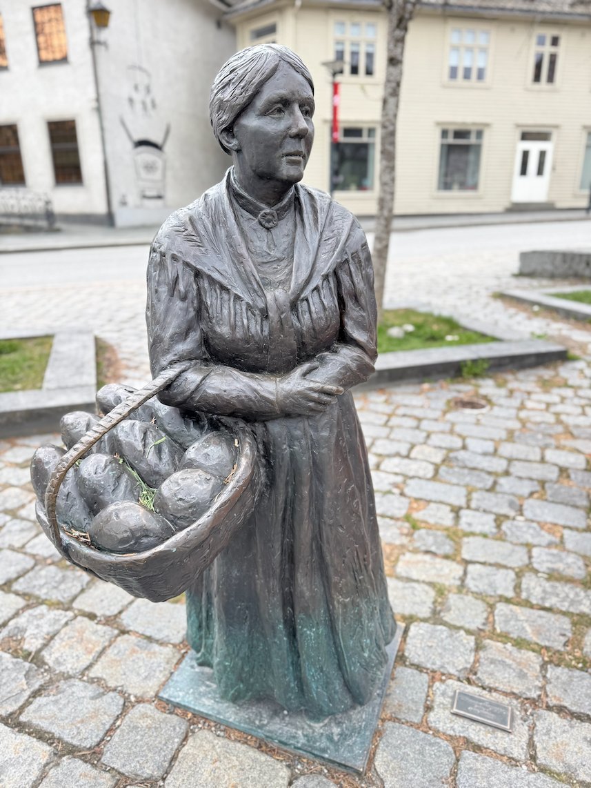 Bronze statue of a woman with a basket on a cobbled square in Voss
