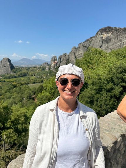 Zayera Khan in a white jacket and sunglasses at Meteora, with rocks and trees behind