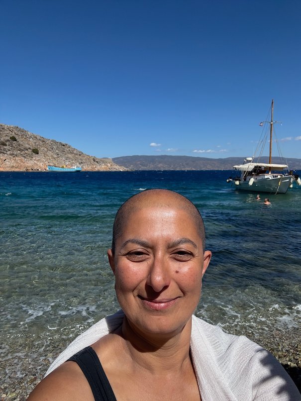 Selfie of Zayera Khan on a beach in Hydra, with a sailing boat in the bay