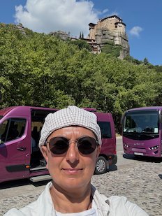 Selfie of Zayera Khan in a cap and sunglasses at Meteora, with a monastery on the hill behind