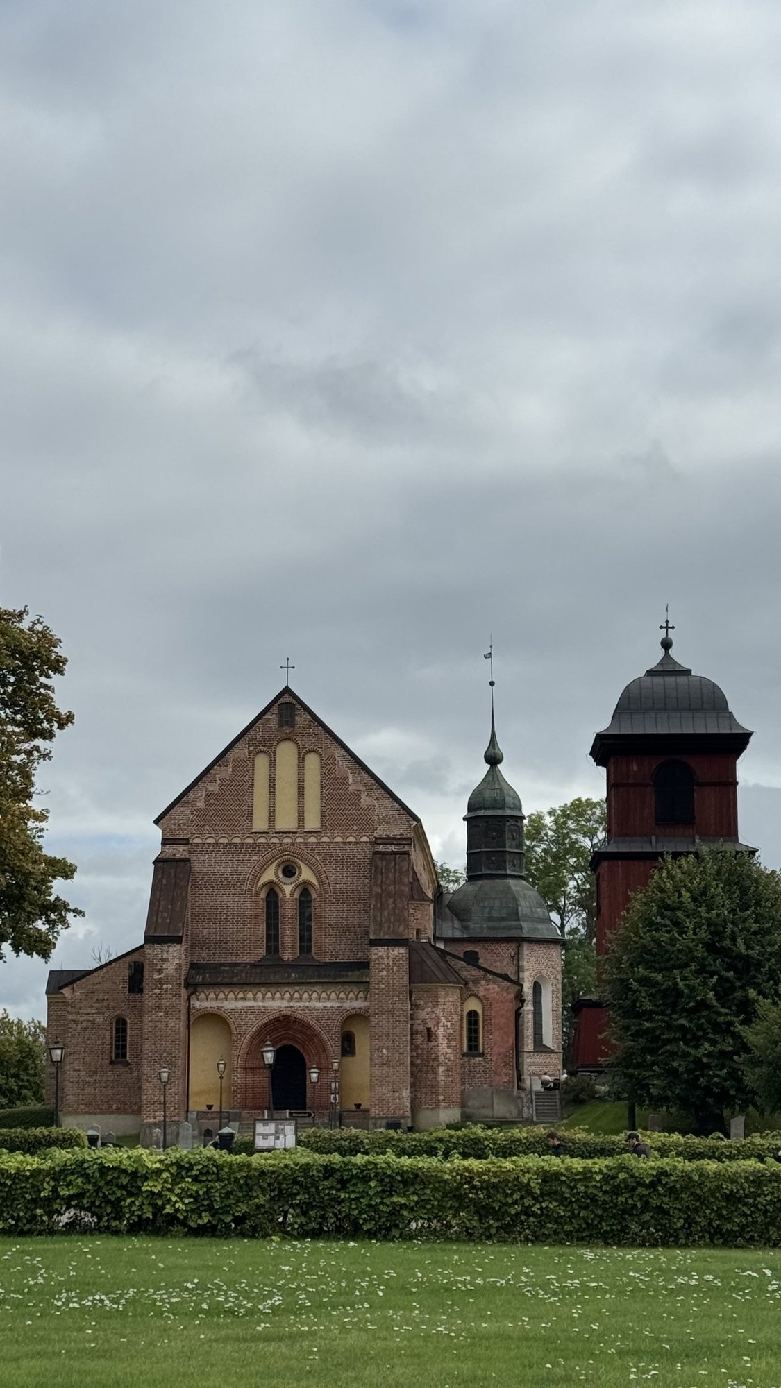 Skokloster Church, a medieval red brick church with a stepped gable and green onion-domed bell tower, next to a separate dark red wooden bell tower with a black domed roof.