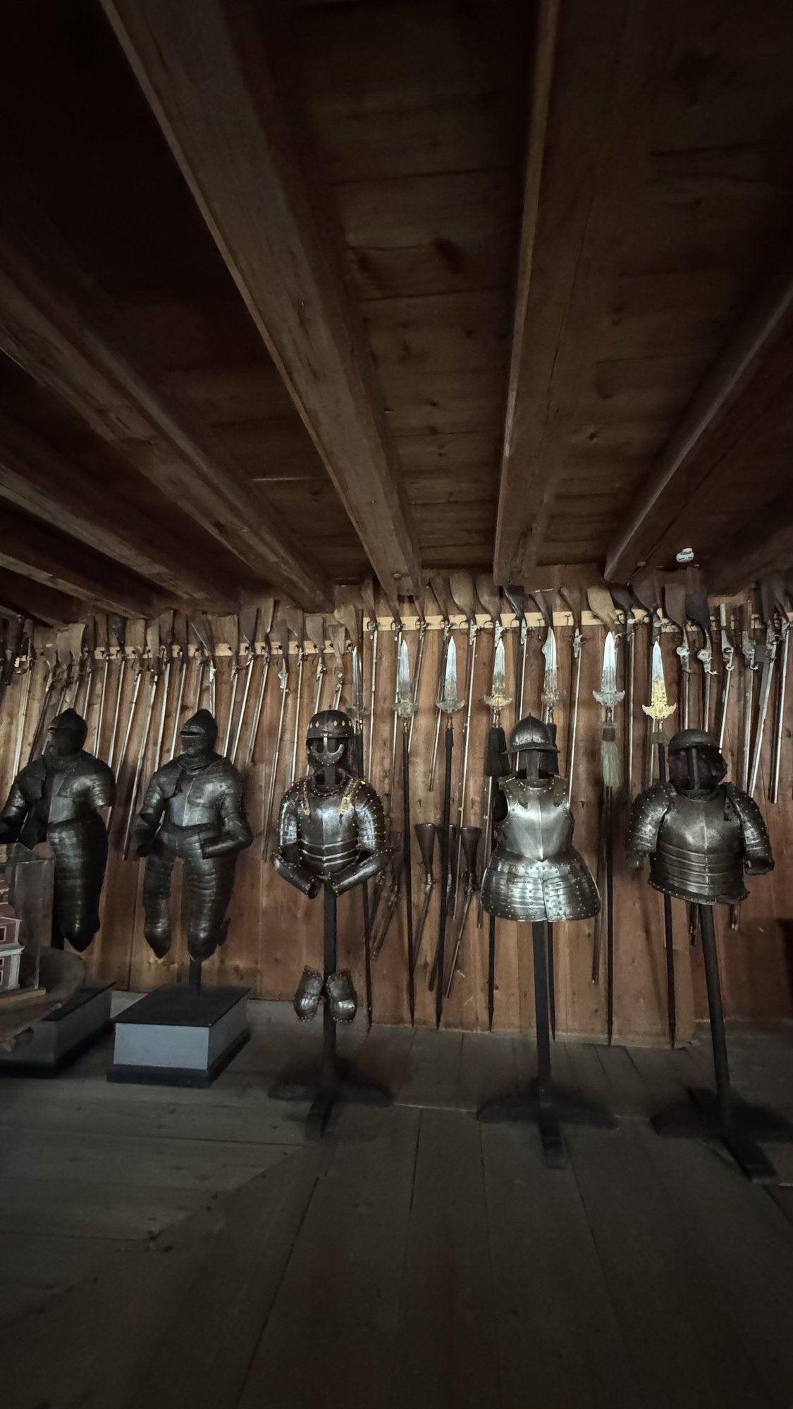 A wood-paneled armory room at Skokloster Castle with swords, pikes, and firearms mounted on the walls, and five suits of armor displayed on stands in a row.
