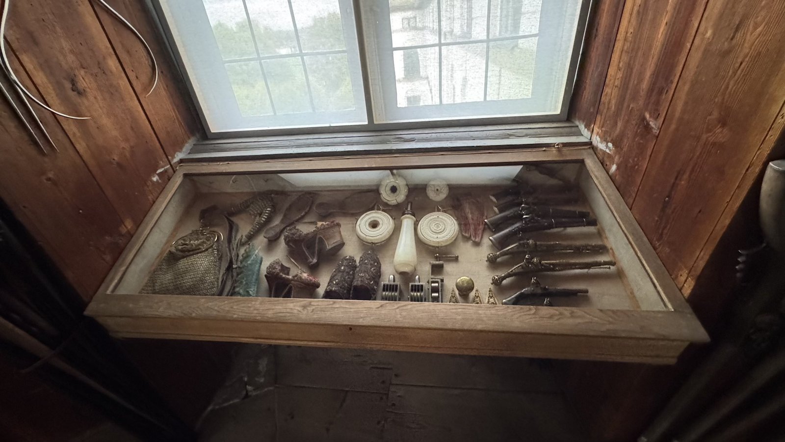 A glass display case built into a window recess at Skokloster Castle, holding small 17th-century curiosities including pistols, powder flasks, a pouch, gloves, and carved objects.
