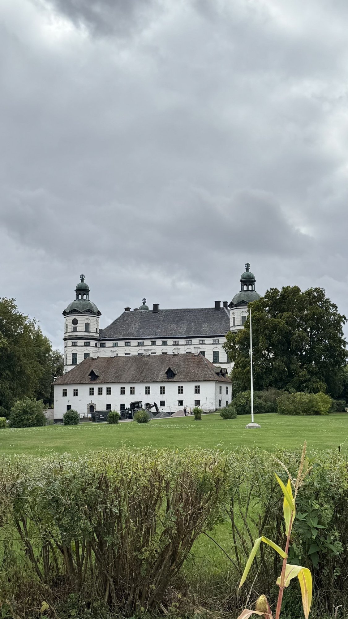 Skokloster Castle, a white baroque building with two green-domed towers, seen across a lawn and hedge under a cloudy sky.