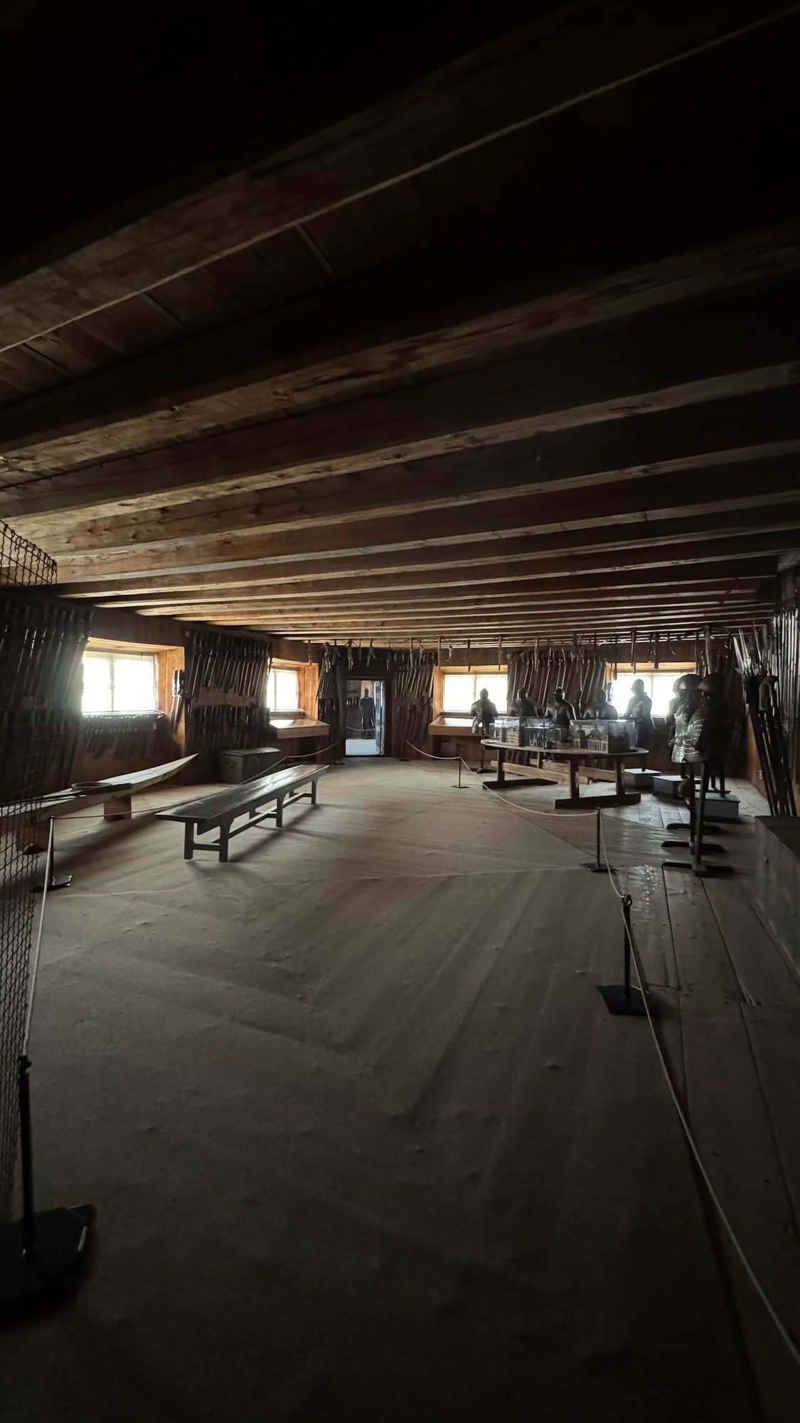 A long room in Wrangel's Armoury at Skokloster Castle with a wood-beamed ceiling, weapon racks along both walls, and suits of armor grouped at the far end.