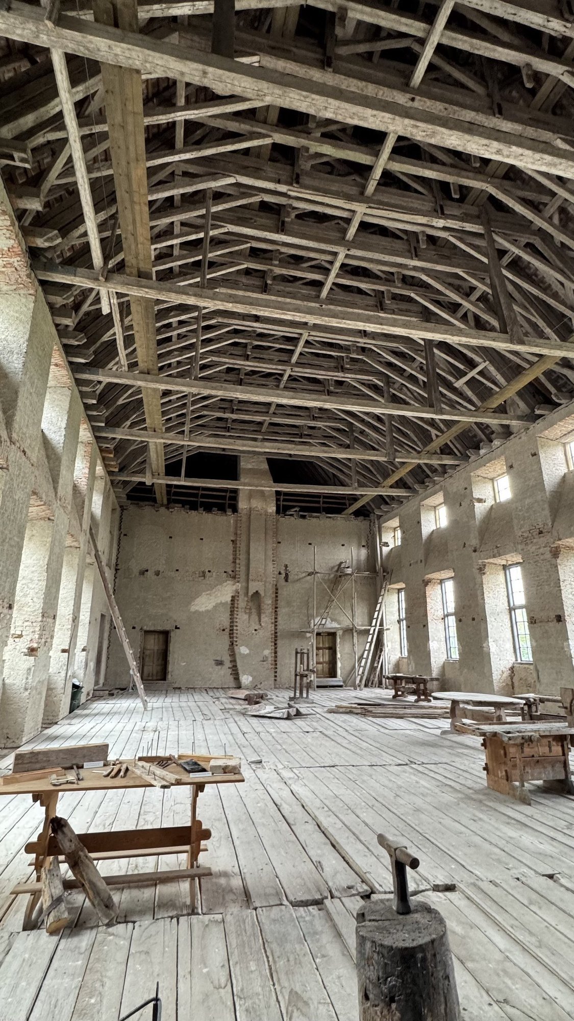 The Unfinished Hall at Skokloster Castle: a tall room with exposed wooden roof trusses, unplastered brick walls, and builders' tools, a sawhorse workbench, and ladders left on the floor.