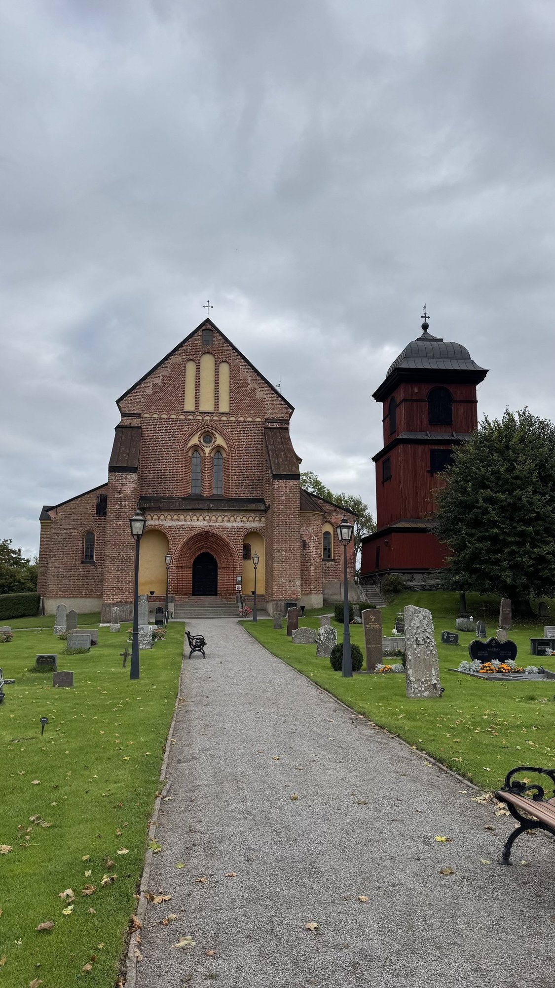 Skokloster Church seen from the cemetery path, with gravestones on both sides, the red brick church ahead, and its wooden bell tower to the right.