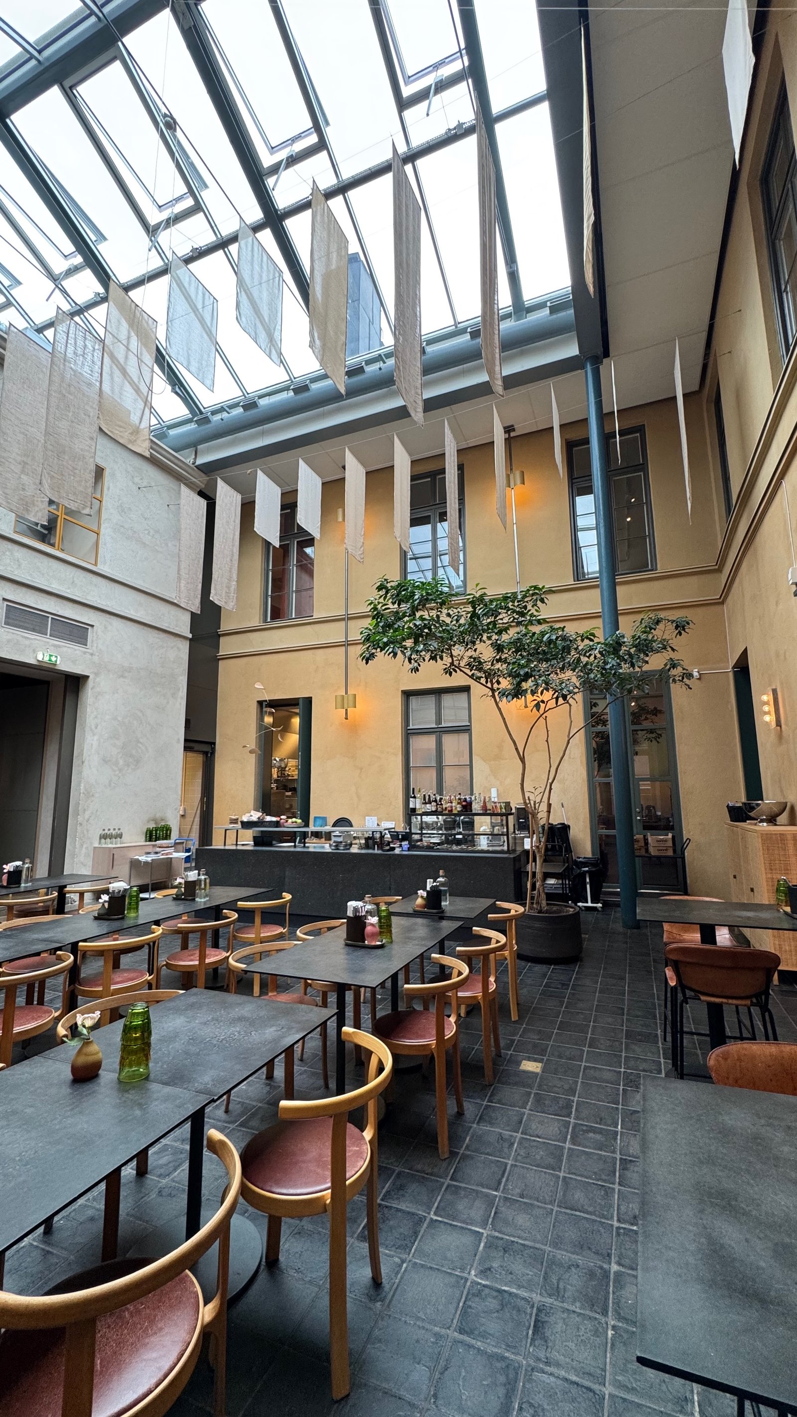 The glass-roofed courtyard of Restaurant AVI, with pale linen banners hanging from the ceiling, wooden tables and chairs, and a small tree growing in the center.