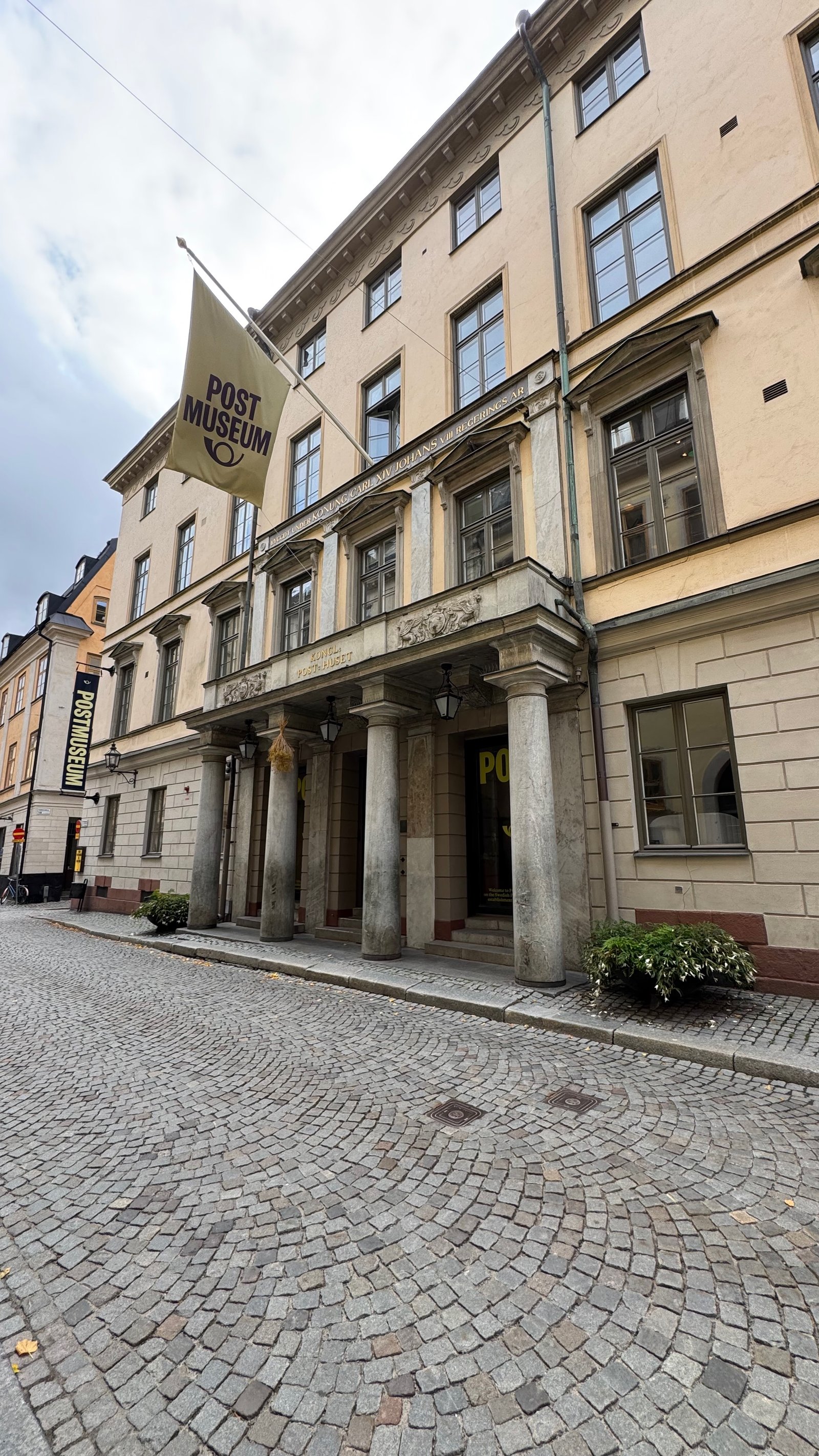 The cream-colored facade of Postmuseum on Lilla Nygatan in Stockholm, with a stone-columned entrance and a golden POSTMUSEUM flag hanging above the door.