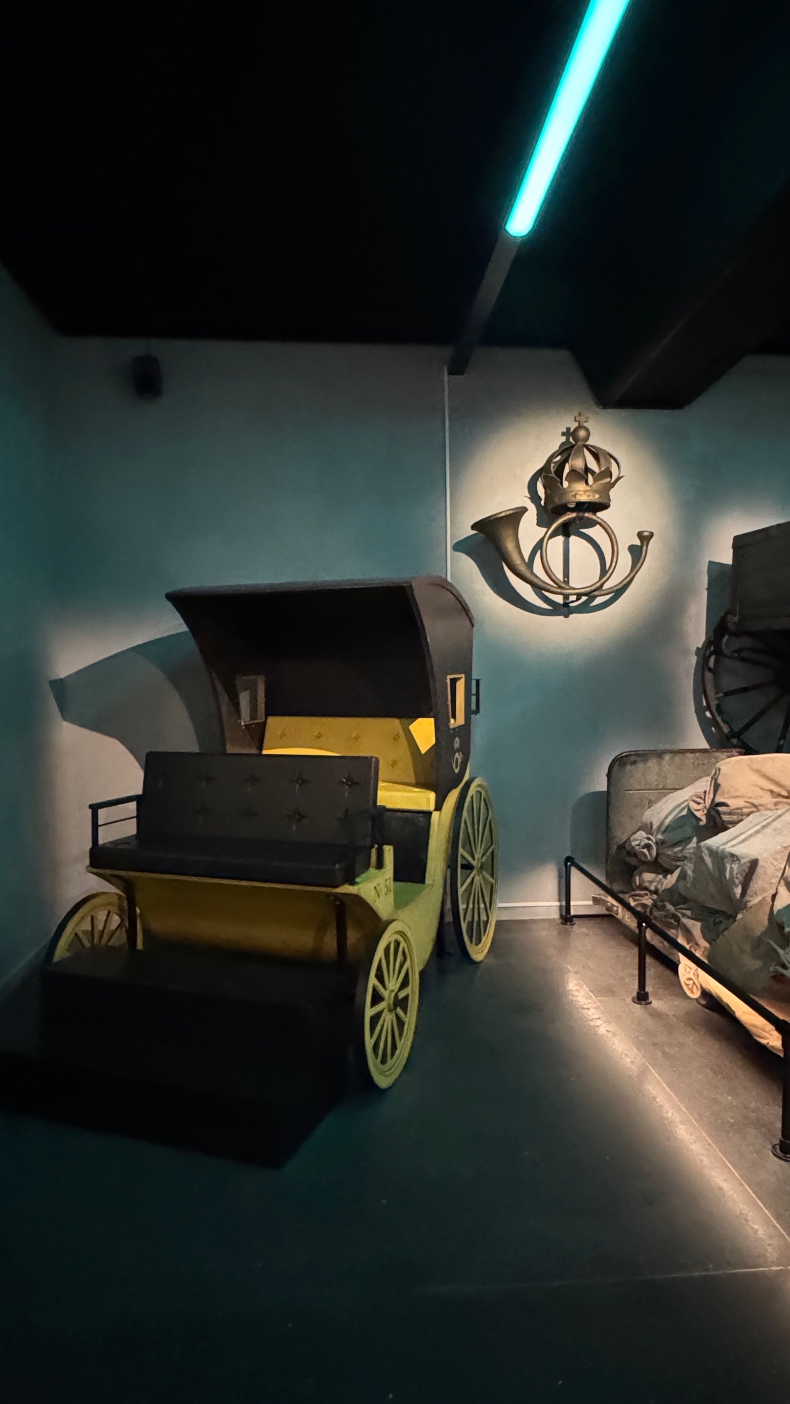 A yellow and black 18th-century mail carriage on large wooden wheels, standing below a gilded royal crown-and-posthorn wall emblem.