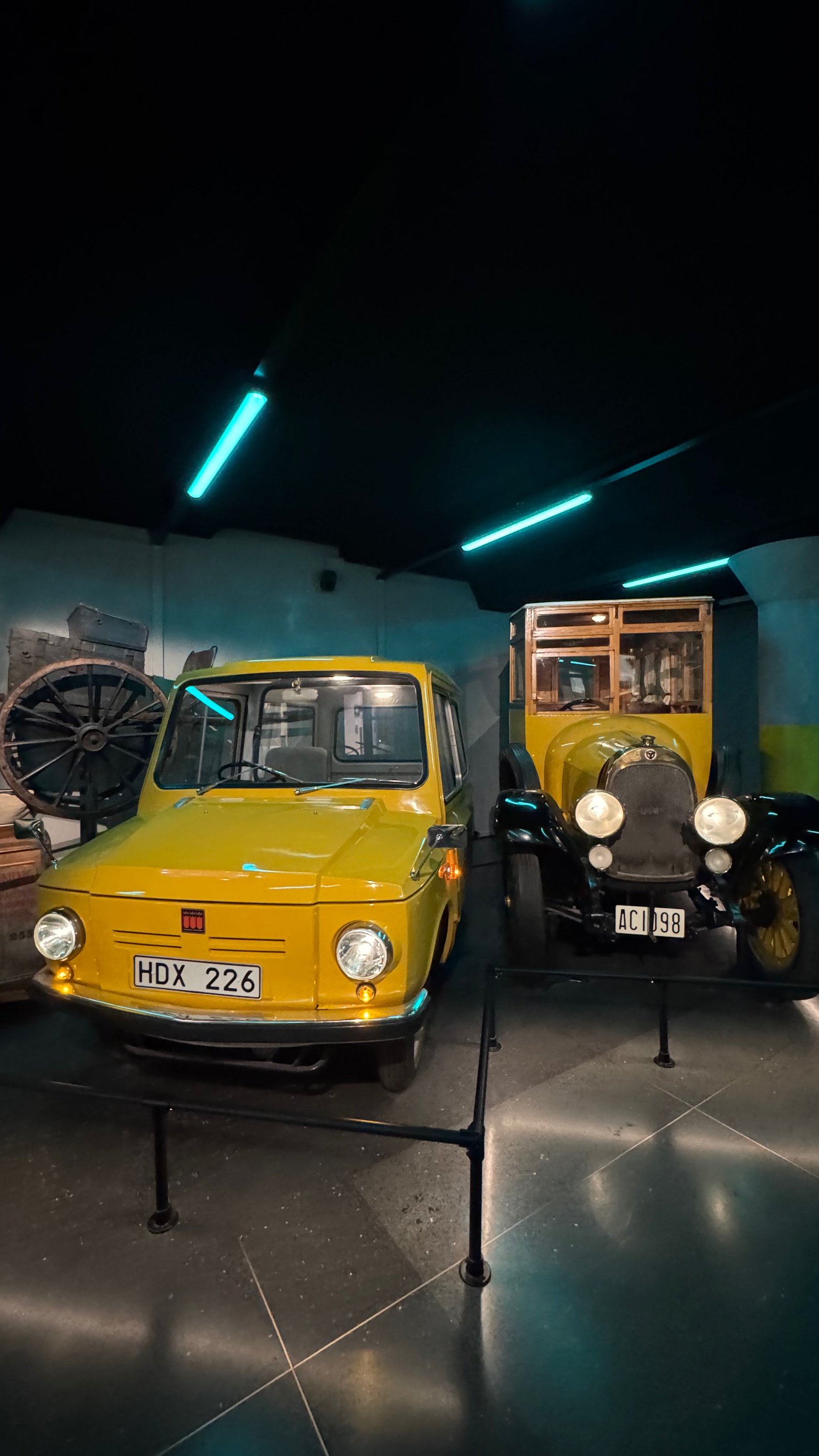 A bright yellow postal delivery van from the 1970s next to an antique black postal carriage-car, displayed inside the museum under blue lighting.