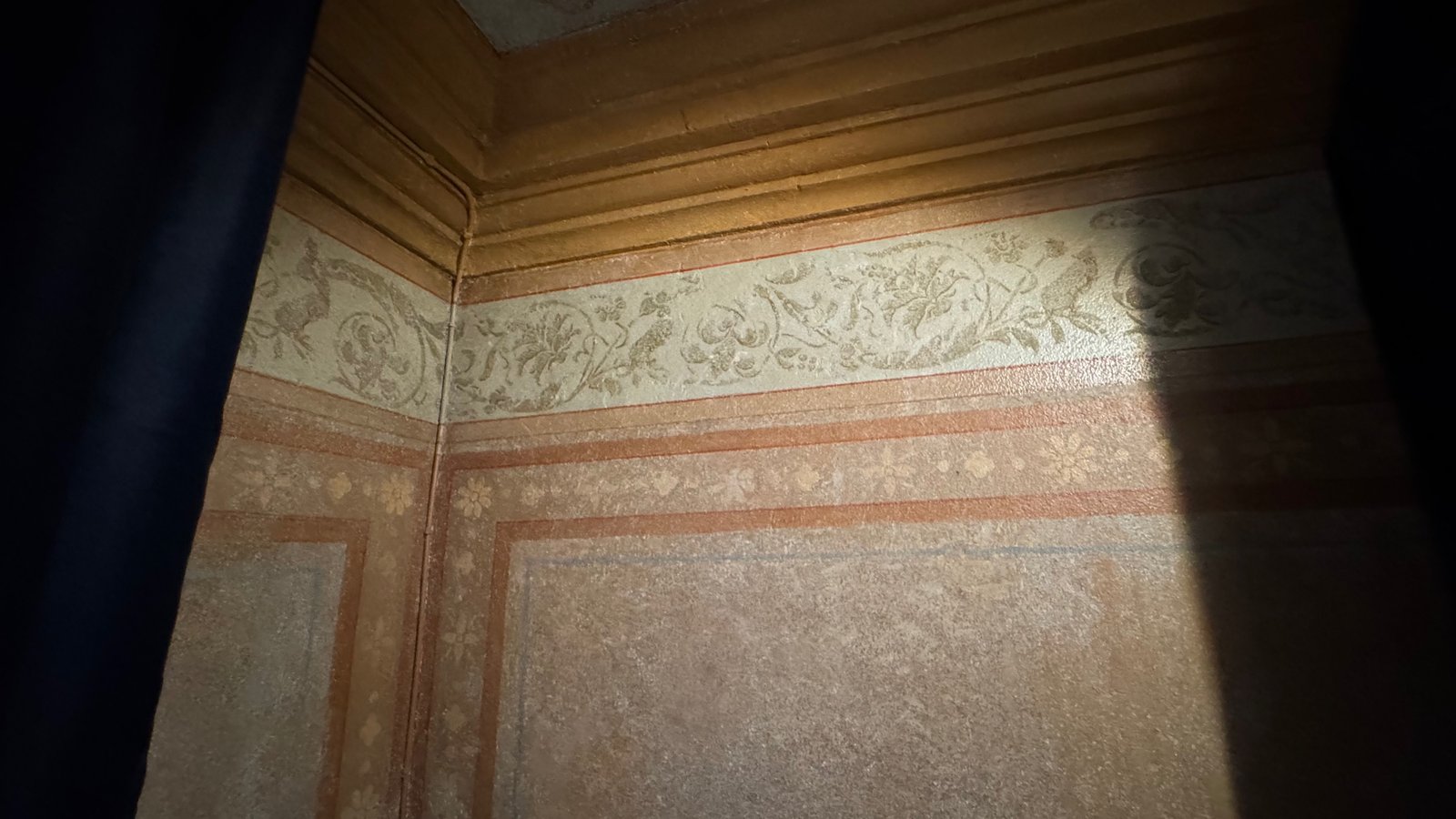 Original 19th-century wall paintings in soft pink, gold, and cream, with floral and bird motifs along the top border, lit by daylight from a window.