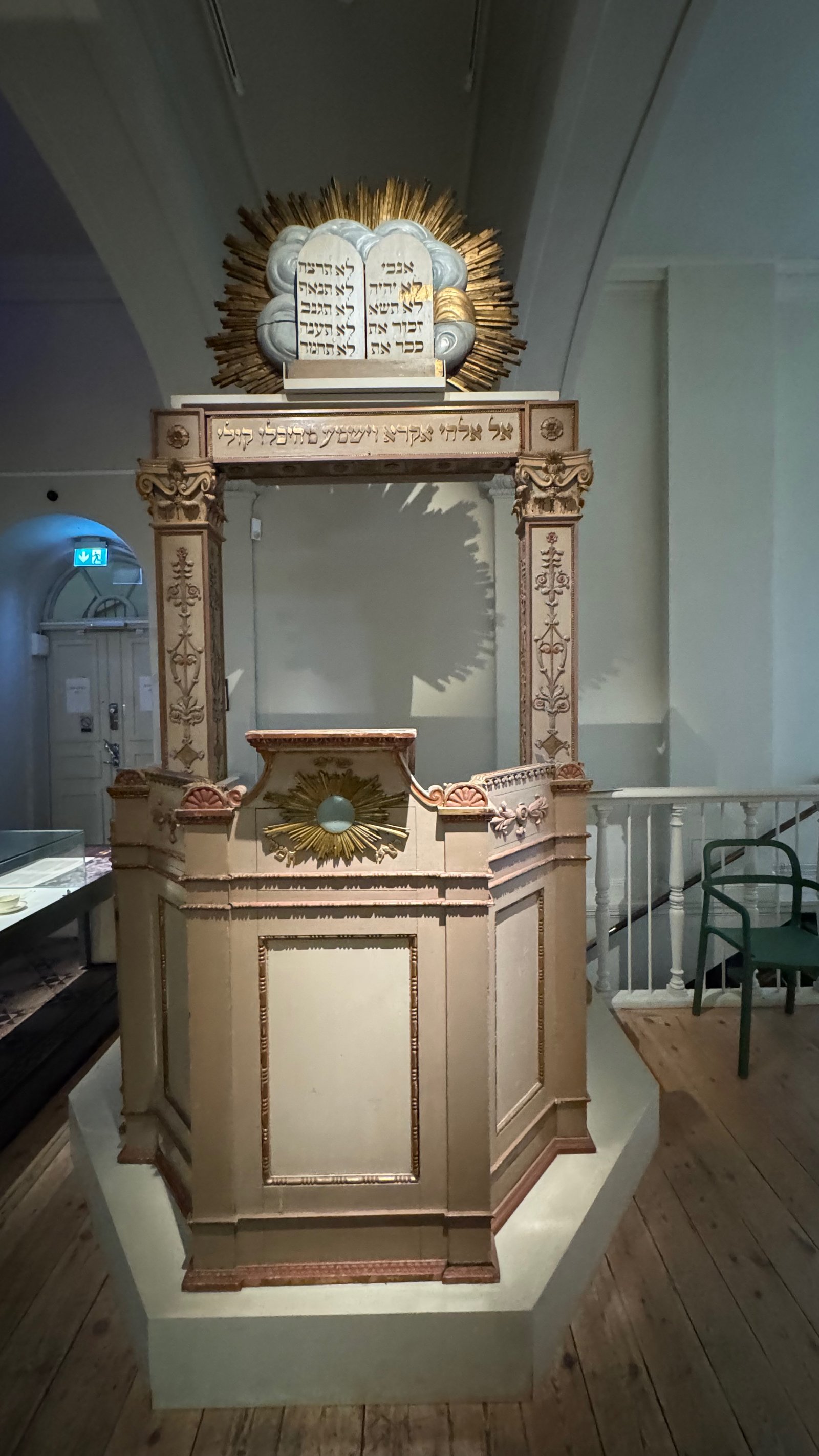 Wooden Torah ark from a synagogue, painted in soft pink and gold, topped with a gilded relief of the Ten Commandments tablets and Hebrew text, on display inside the museum.