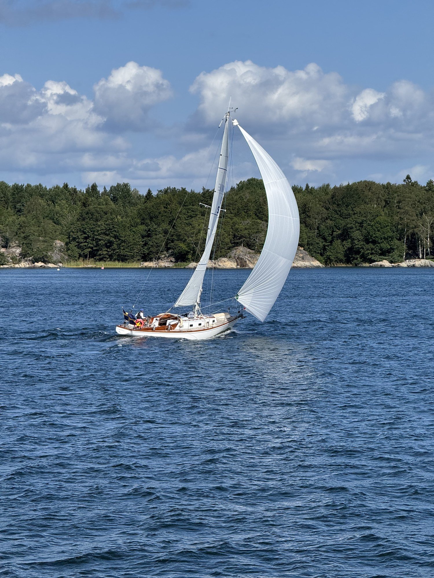 A sailboat with white sails on blue water near Möja, with a green forest shore behind it.