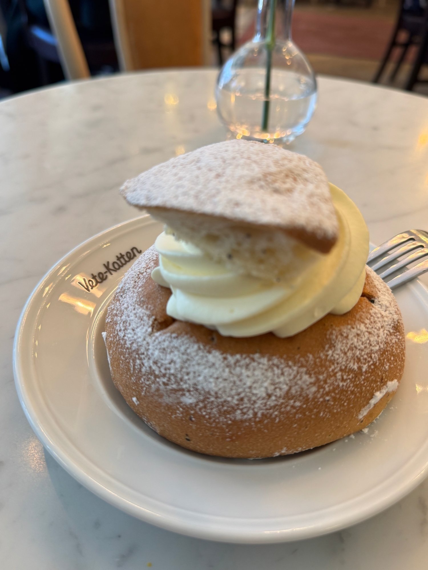 A semla, a cardamom bun filled with whipped cream and dusted with powdered sugar, on a plate on a marble café table.