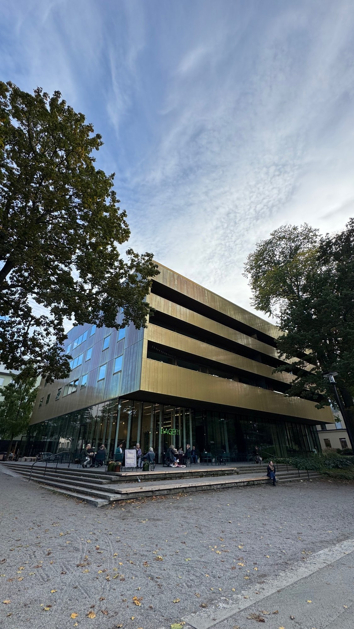 Gold-colored facade of Sven-Harrys Konstmuseum with large trees on both sides and people in front of the glass ground floor.