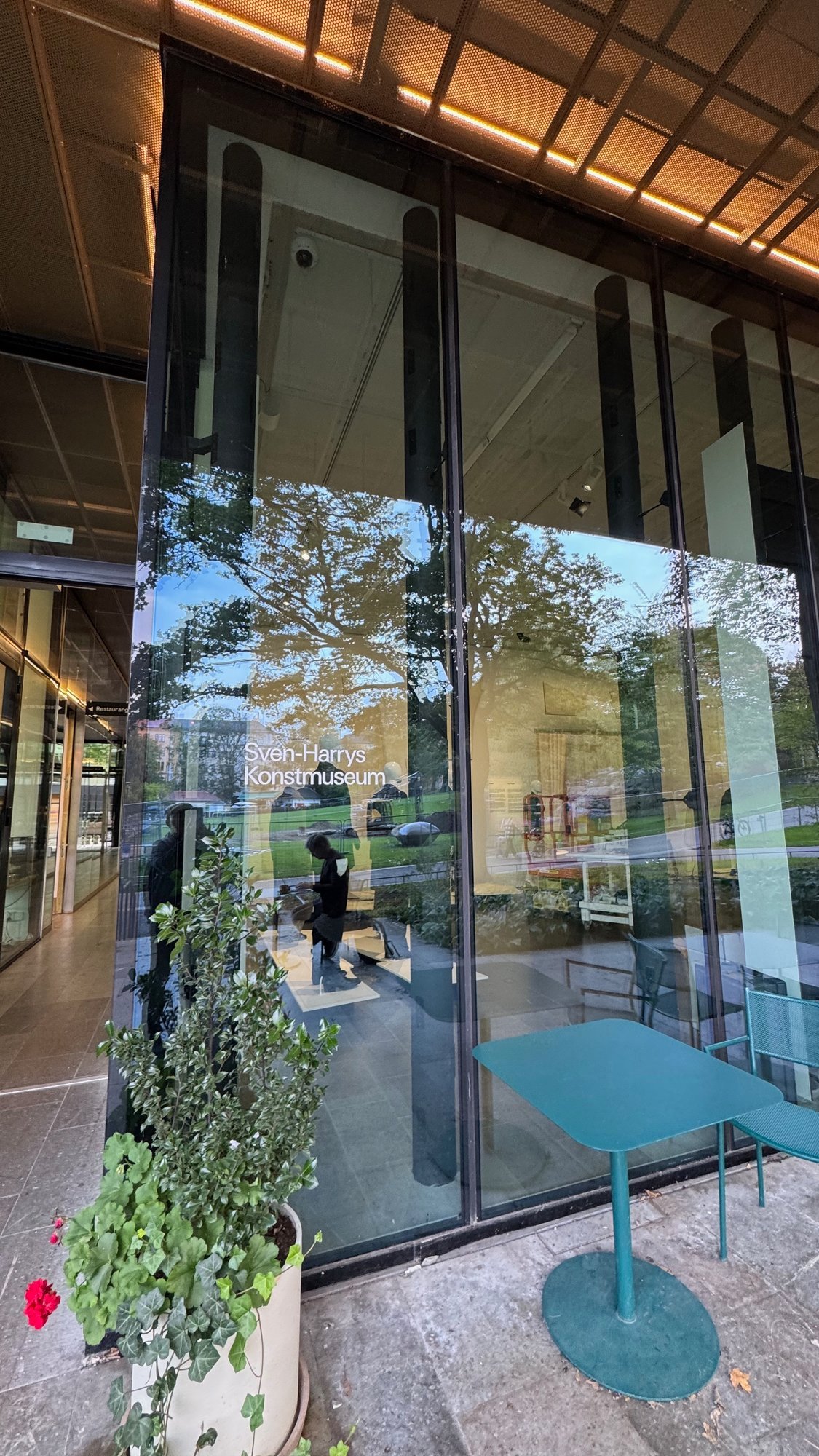 Glass front of Sven-Harrys Konstmuseum with the museum name on the window. A teal outdoor table and a potted plant stand in front.